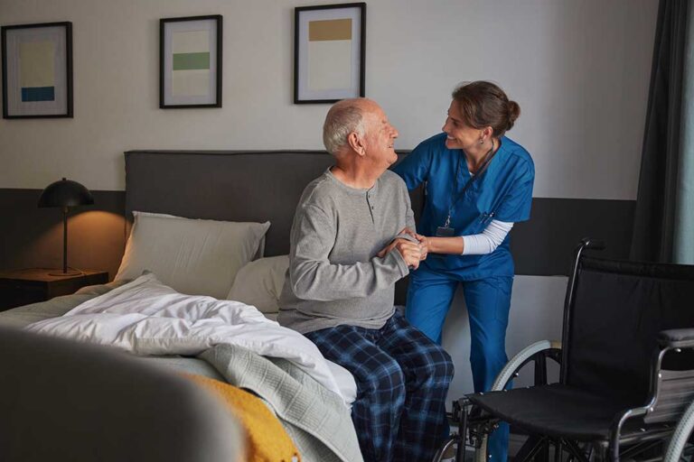 Two seniors chat at a dining table with a caregiver assisting gently in a bright communal room.
